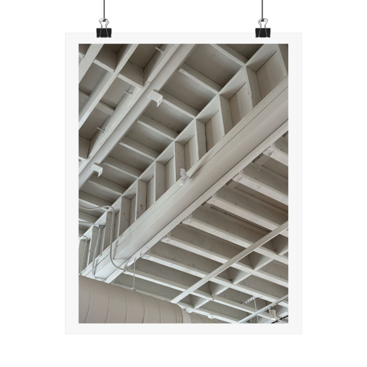 Print of a photograph looking up at white painted ceiling beams and pipework, shown hanging from binder clips.