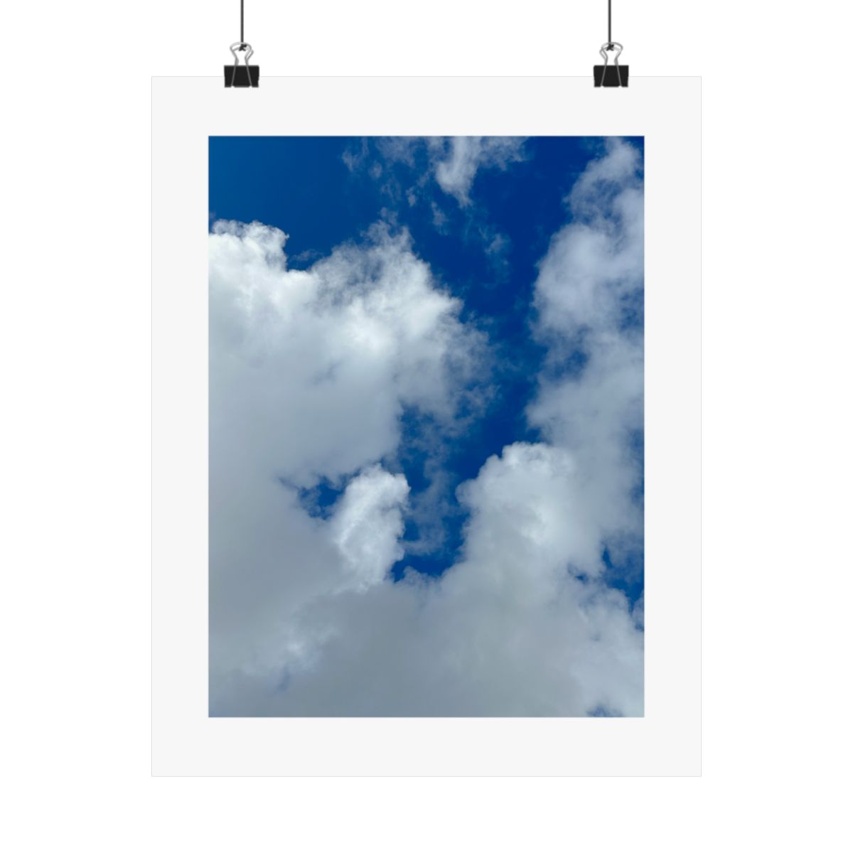 Print of a photograph looking up at bright white clouds against deep blue sky, shown hanging from binder clips.