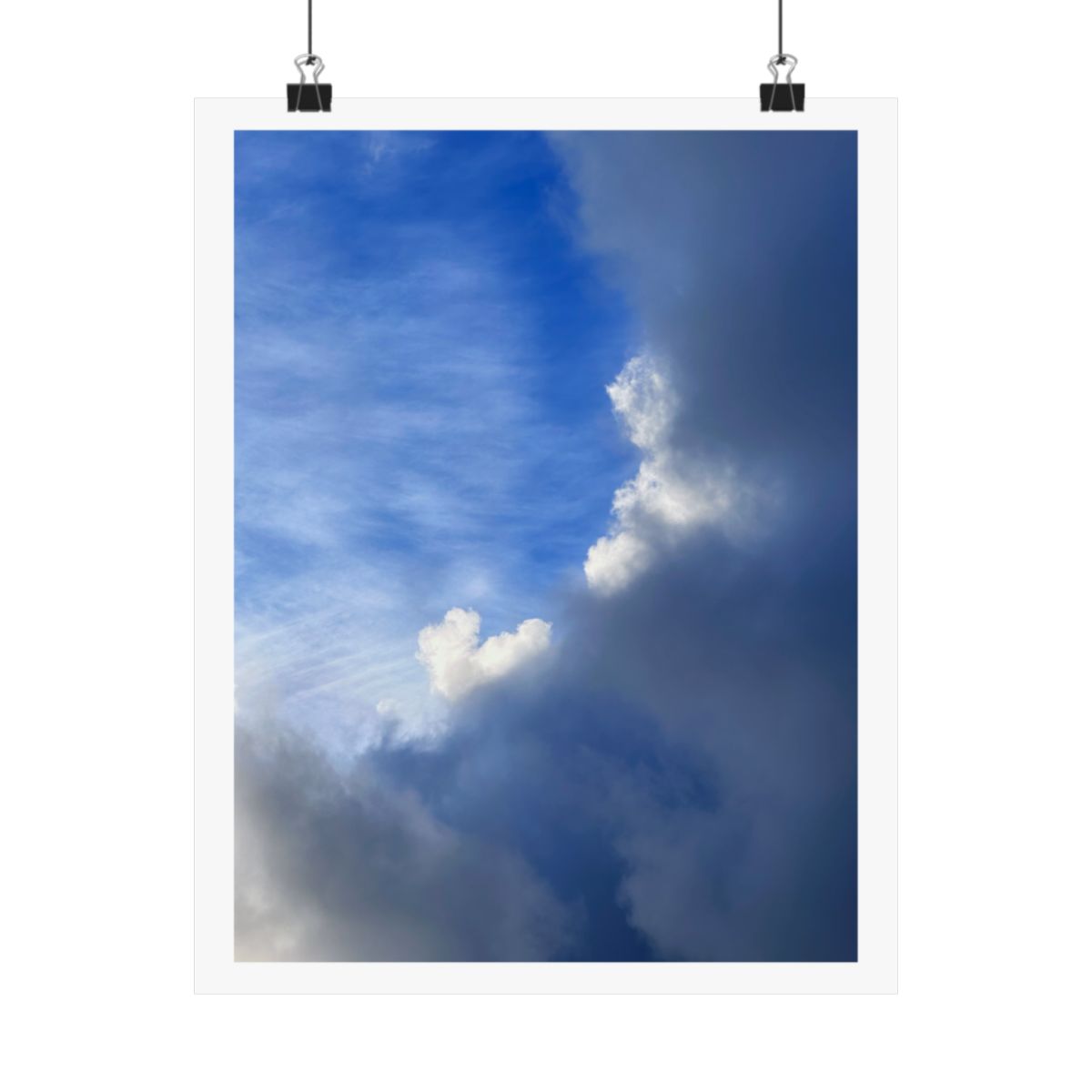 Print of a photograph of a tall white cloud rising through grey haze against dark blue sky, shown hanging from binder clips.