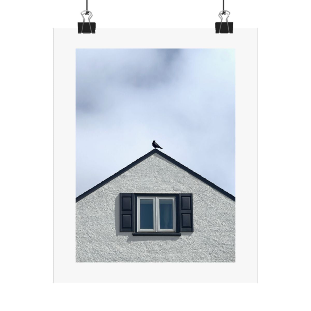 Print of a photograph of a crow on the peak of a pale gabled roof above a dark window, shown hanging from binder clips.