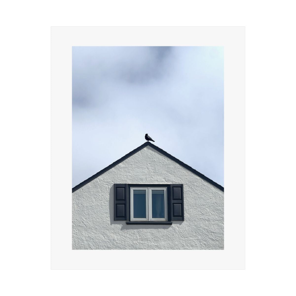 Print of a photograph of a crow on the peak of a pale gabled roof above a dark window.