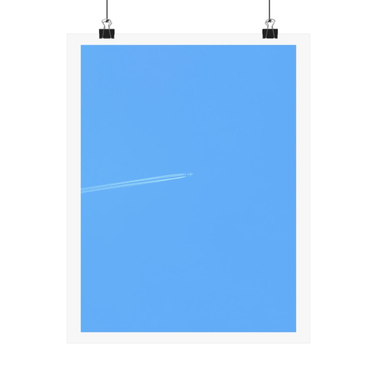 Print of a photograph of pale blue sky crossed by a single faint aeroplane contrail, shown hanging from binder clips.