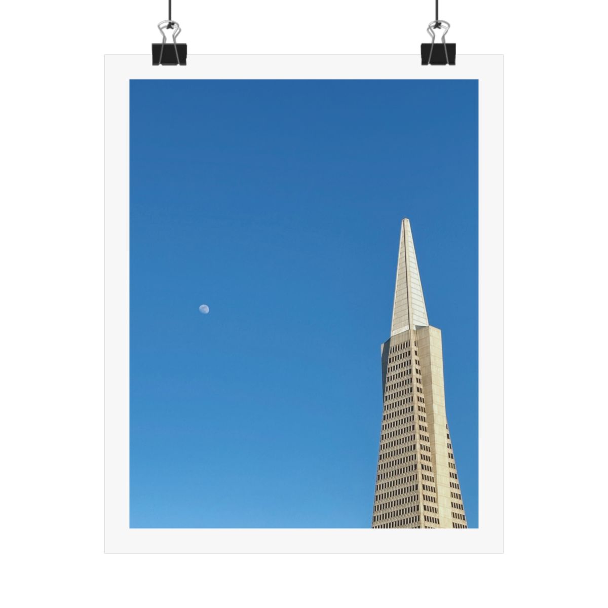 Print of a photograph of the Transamerica Pyramid against deep blue sky with a pale moon, shown hanging from binder clips.
