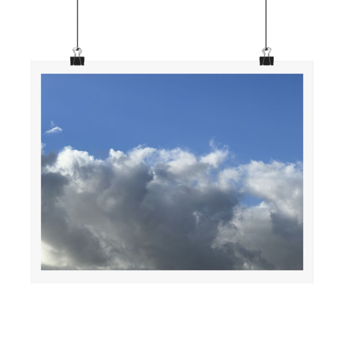 Print of a photograph of a wide bank of white and grey cloud beneath open blue sky, shown hanging from binder clips.