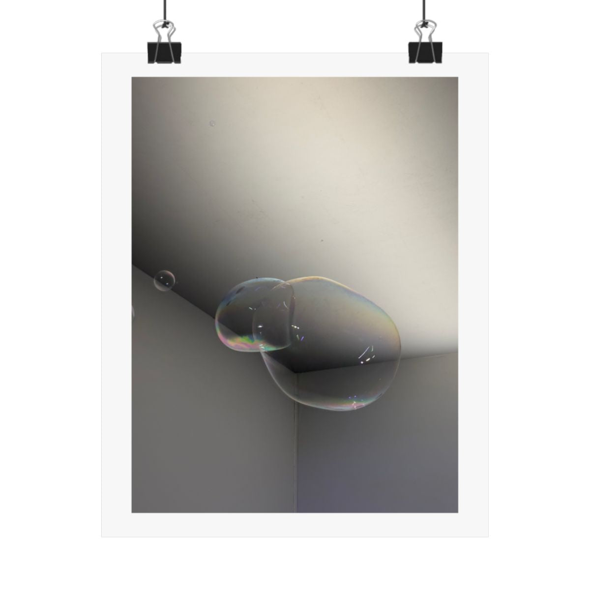Print of a photograph of a single soap bubble in the corner of a pale empty room, shown hanging from binder clips.
